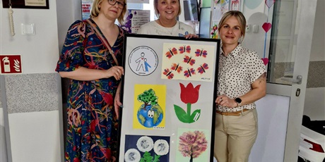 Powiększ grafikę: obok siebie stoją trzy kobiety,  jedna z nich która stoi w środku trzyma w ręku ramę z pracami plastycznymi dzieci. 