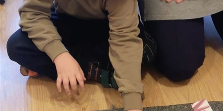 Powiększ grafikę: Chłopiec i pani pracują na dywanie Montessori na podłodze. Chłopiec składa pocięty obrazek buraka z literami, tak żeby powstał wyraz burak i jego zdjęcie.