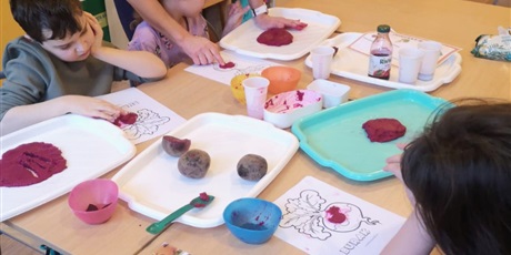 Powiększ grafikę: Trójka dzieci bawi się przy stolikach masą sensoryczną z buraka. Na stole przepołowione buraki.