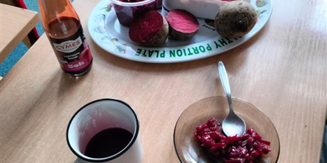 Powiększ grafikę: Stół gdzie na talerzyku leży cały burak, przekrojone buraczki i masa sensoryczna z buraków. Obok stoi buteka z sokiem, kubek z sokiem z buraków i surówka z buraczków.