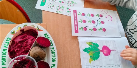 Powiększ grafikę: Chłopiec robi katy pracy w zeszycie tematycznie związane z burakiem. Obok talerz z masą sensoryczną i buraczkami.