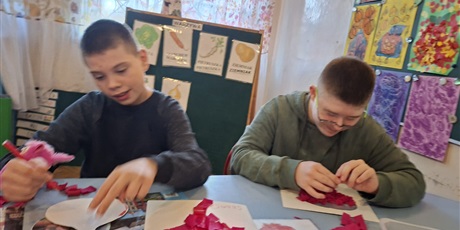 Powiększ grafikę: Dwóch chłopców przy stole wykonują pracę plastyczną z buraczka. Jeden rwie bibułę i wykleja kontur buraka. Drugi obrysowuje kontur mazakiem.