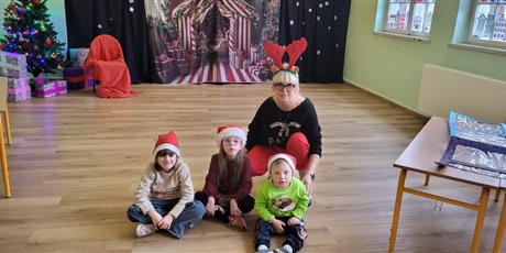 Powiększ grafikę: Dziewczynki i mały chłopiec siedzą obok swojej wychowawczyni. W tle widać śiteczne tło oraz chionkę.