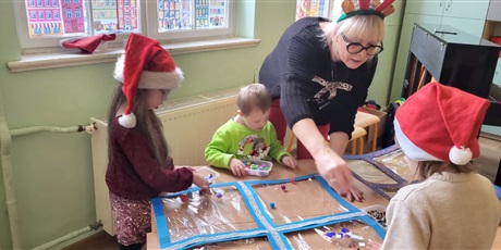 Powiększ grafikę: Dzieci wykonują prace plastyczną. Wyklejają ozdobami wielki prezent. Pomaga im nauczycielka.