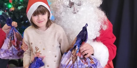 Powiększ grafikę: ziewczynka stoi obok Mikołaja. Mikołaj wręcza jej prezent. W tle choinka.