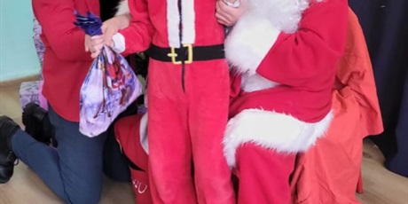 Powiększ grafikę: Chłopiec stoi obok Mikołaja. Mikołaj wręcza prezenty. W tle choinka.