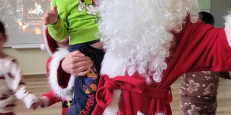 Powiększ grafikę: Mikołaj, trzaymając chłopca na rękach, tańczy. Dookoła Mikołaja tańczą dzieci w kole.