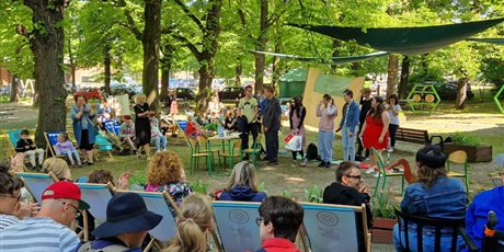 Powiększ grafikę: Widzowie siedzą tyłem do kamery, przed nimi artyści teatralni przedstawiają spektakl, w tle park sensoryczny i drzewa