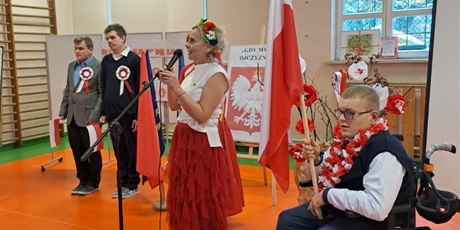 Powiększ grafikę: Nauczycielka w stroju w kolorach flagi Polski stoi obok uczniów z proporcami biało-czerwonymi. W tle tablice z materiałami dotyczącymi apelu.