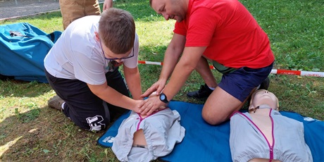 Powiększ grafikę: ratownik medyczny uczy chłopca resuscytacji na manekinie. Na drugim planie zieleń.