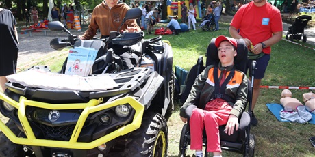 Powiększ grafikę: Chłopiec siedzący na quadzie obok Niego chłopiec na wózku inwalidzkim oraz ratownik . W tle grupa bawiących się dzieci w plenerze 