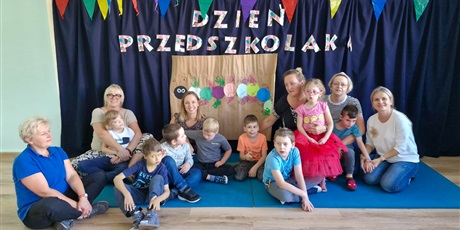 Powiększ grafikę: Nauczycielki i dzieci siedzą na podłodze. Za nimi napisem na ścianie "dzień przedszkolaka".