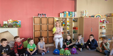 Powiększ grafikę: 3.	Nauczycielka siedzi na krzesełku, wokół niej siedzą dzieci. W tle meble