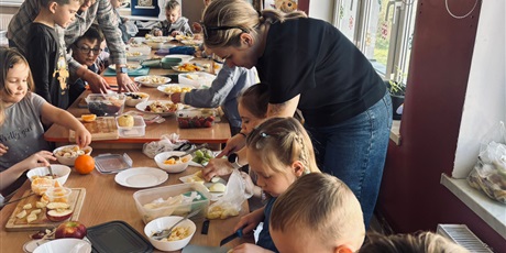 Powiększ grafikę: 2.	Dzieci siedzą przy stoliku a panie pomagają im w krojeniu owoców potrzebnych do przygotowania sałatki.