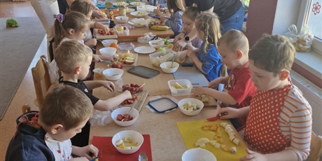 Powiększ grafikę: 7.	Dzieci siedzą przy stole a nad nimi pochylając się nauczycielki, które im pomagają.