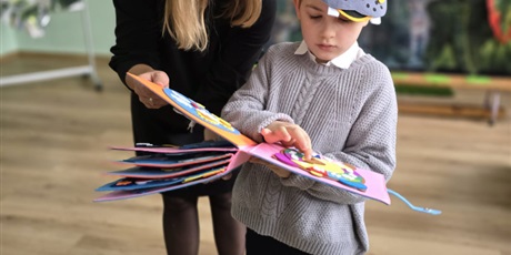 Powiększ grafikę: Uczeń i nauczycielka przeglądają książkę kolorową. W tle wystrój na pasowanie.