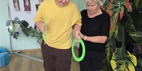 Powiększ grafikę: Wychowanek i nauczyciel rzucają kółkami do celu. W tle Dekoracja i tablica ze zdjęciami nowoprzyjętych przedszkolaków.