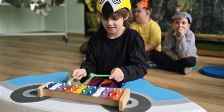 Powiększ grafikę: Wychowanka gra na cymbałkach. W tle dzieci siedzące na podłodze i dekoracja na pasowanie.