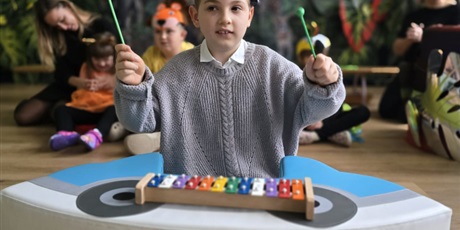 Powiększ grafikę: Wychowanek gra na cymbałkach. W tle nauczyciel i dzieci siedzące na podłodze i dekoracja na pasowanie.