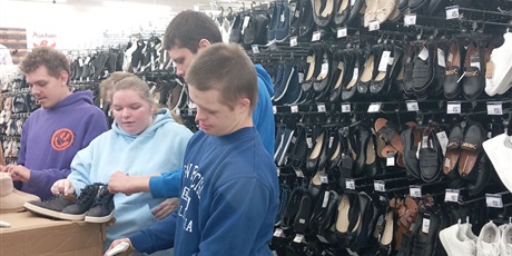 Powiększ grafikę: Uczniowie zakładają zabezpieczenia antykradzieżowe na obuwie. W tle wieszaki z butami