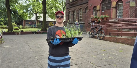 Powiększ grafikę: 10.	Uczeń niesie rozsady roślin. W tle budynek szkoły