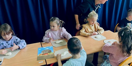 Powiększ grafikę: 2.	Dzieci siedzą przy stoliku ,za nimi stoi nauczycielka nadzorując prace dzieci.