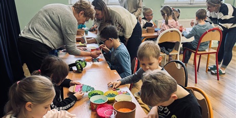 Powiększ grafikę: 8.	Dzieci siedzą przy stoliku a nad nimi pochylając się panie, które pomagają im przygotować jajka wielkanocne.