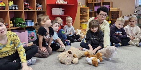 Powiększ grafikę: 3.	Dziewczynka siedzi na podłodze a przed nią ułożone są misie. Obok siedzą inne dzieci.