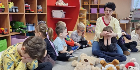 Powiększ grafikę: 5.	Chłopiec siedzi na podłodze a przed nim ułożone są misie. Obok siedzą dzieci.