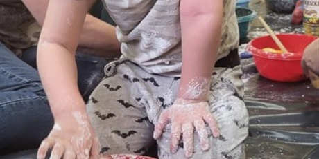 Powiększ grafikę: Chłopiec siedzi z nauczycielem na zabezpieczonej folią malarską podłodze. Przed sobą ma miskę, w której, powstaje masa z mąki ziemniaczanej i oleju rzepakowego tzw. piasek kinetyczny.
