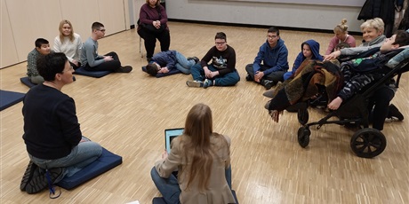 Powiększ grafikę: Siedzą uczniowie i nauczyciele w kręgu na podłodze w dużej sali. Jedna z nauczycielek trzyma otwarty laptop.