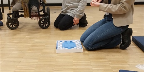 Powiększ grafikę: Siedzą dwie kobiety na podłodze obok jednej z nich siedzi chłopiec na wózku. Jedna z nich prezentuje małe flakoniki z zapachami.