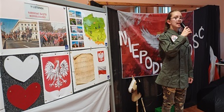 Powiększ grafikę: Uczennica w stroju zołnierza śpiewa piosenkę do mikrofonu. W tle tablica tematyczna i symbol orła białego z napisem NIEPODLEGŁOŚĆ.