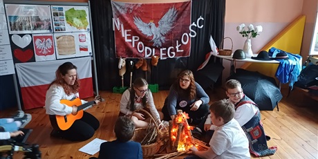 Powiększ grafikę: Dzieci siedzące wokół sztucznego ogniska . Obok kosz wiklinowy z gałęziami. Nauczycielka siedzi przy dzieciach  i gra na gitarze. W tle tablice i scenografia oraz różne rekwizyty patriotyczne.  
