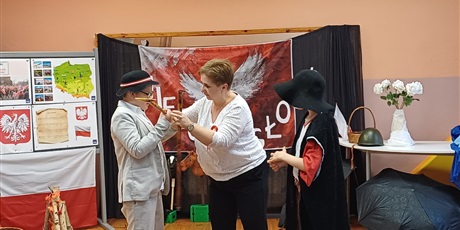 Powiększ grafikę: Nauczycielka podaje chłopcu ciupagę do tańca. Na środku sali stoi dwóch chłopców w góralskich kapeluszach. W tle dekoracja okolicznościowa- Święto Odzyskania Niepodległości. 