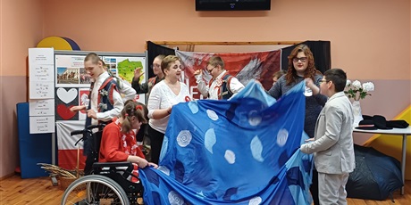 Powiększ grafikę: Dzieci z nauczycielką trzymają niebieska płachtę ( Wisła ). Wachlują materiałem. W tle dzieci tańcżace krakowiaka w regionalnych strojach. 