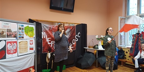 Powiększ grafikę: Dziewczynka śpiewająca do mikrofonu. Obok uczennica przebrana za żołnierza machająca flagą Polski. W tle dekoracja. 