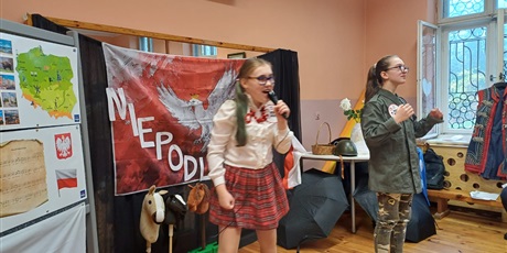 Powiększ grafikę: Dziewczynka w stroju krakowianki śpiewająca do mikrofonu. Koleżanka w stroju żołnierza pokazuje gesty do piosenki. 