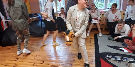 Powiększ grafikę: Dzieci biegające w koło z końmi na kijach. W tle uczniowie i nauczycielka siedzący na ławce. 