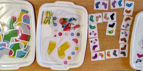 Powiększ grafikę: Na stole leżą tace z zalaminowymi kolorowymi skarpetkami do dobierania w pary, tacę z skarpetami i klamerkami do wyboru kolorów, Domino skarpetkowe.