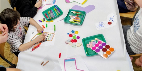 Powiększ grafikę: Zdjęcie zrobione z góry, dzieci na warsztatch siedzą przy złączonych stołach i ozdabiają skarpetki. Na stole leżą mazaki, kredki, elementy ozdobne.