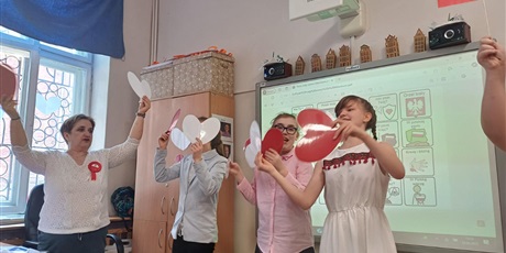 Powiększ grafikę: Trójka dzieci i nauczycielka pokazują białe i czerwone serduszka. W tle tablica multimedialna