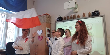 Powiększ grafikę: Chłopiec z dużą biało-czerwoną flagą w ręku. Po obu jego stronach nauvzycielki z przypiętymi kotylionami. W tle dwie dziewczynki i tablica multimedialna. 