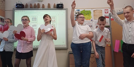 Powiększ grafikę: Trzy dziewczynki i tzech chłopców. Dziewczynki trzymają białe i czerwone serca. Chłopcy- flagi. W tle tablica multimedialna i dekoracja przedstawiająca mapę Polski, hymn, godło i flagę.