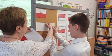 Powiększ grafikę: Przy tablicy chłopiec i nauczycielka. Wspólnie przypinają białą kartkę, nad którą wisi wzór flagi Polski. Obok flagi biało-czerwone w różnych kombinacjach.