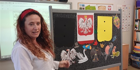 Powiększ grafikę: Nauczycielka przy tablicy, na której przyczepione są tła żółte i czerwone, różne ptaki i nakrycia głowy. Wisi też godło Polski