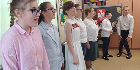 Powiększ grafikę: Siedmioro dzieci i nauczycielka stoją odświętnie ubrani. W tle tablica z symbolami patriotycznymi.