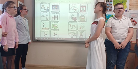 Powiększ grafikę: Po lewej stronie tablicy dwie dziewczynki, po prawej dziewczynka i chłopiec. Na tablicy multimedialnej wiersz "Kto ty jesteś" z symbolami AAC i tekstem.