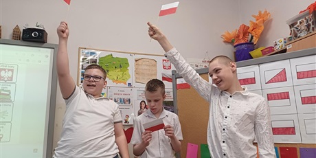Powiększ grafikę: Trzech chłopców w białych koszulach i flagami Polski w ręku. W tle widoczne dekoracje, tablice tematyczne, symbole narodowe.