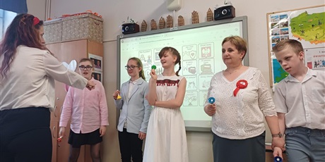 Powiększ grafikę: Trzy dziewczynki, nauczycielka i chłopiec stoją przed tablicą multimedialną. W rękach trzymają kolorowe dzwonki. Przed nimi stoi nauczycielka, która palcem wskazuje na dziewczynkę, która ma grać.